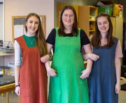 three women stood side by side linking arms with linen aprons on in rust colour brown, bright green and blue